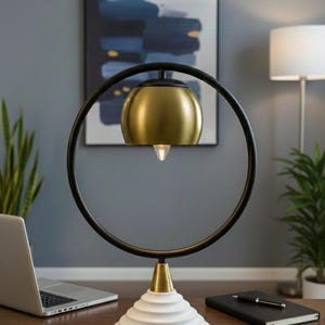 May include: A modern desk lamp with a gold-colored shade and a black circular frame. The lamp sits on a white, cone-shaped base. A laptop, notebook, and pen are on the wooden desk.