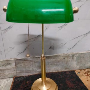 May include: A green-shaded banker's lamp with a brass-colored base and stem. The lamp features a rounded, rectangular shade and a pull chain. The base is circular and tiered. The lamp is designed for desk or table use.