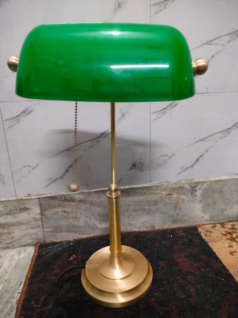 May include: A green-shaded banker's lamp with a brass-colored base and stem. The lamp features a rounded, rectangular shade and a pull chain. The base is circular and tiered. The lamp is designed for desk or table use.