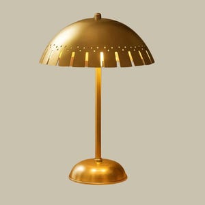 May include: A gold-colored table lamp with a domed shade featuring cut-out slots. The lamp has a slender stem and a rounded base. The design is mid-century modern, with a warm, inviting aesthetic.