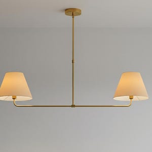 Puede incluir: Una lámpara colgante moderna de dos luces. La lámpara tiene un acabado dorado y presenta dos pantallas beige en forma de cono. La luz cuelga de una barra central y un brazo horizontal, creando un diseño equilibrado.