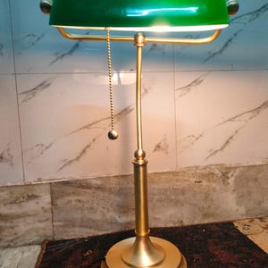 May include: A brass-colored desk lamp with a green shade. The lamp has a rounded base, a slender stem, and a curved shade. A small chain with a ball hangs from the shade. The lamp is lit, casting a warm glow.