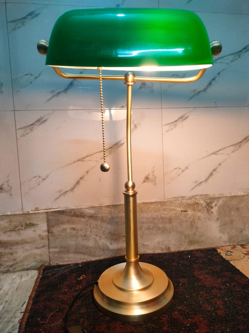 May include: A brass-colored desk lamp with a green shade. The lamp has a rounded base, a slender stem, and a curved shade. A small chain with a ball hangs from the shade. The lamp is lit, casting a warm glow.