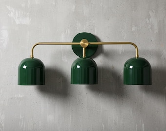 May include: A modern wall-mounted light fixture with three dark green, dome-shaped shades. The shades are suspended from a brass-colored horizontal bar. The fixture is mounted on a circular dark green base with a brass accent.