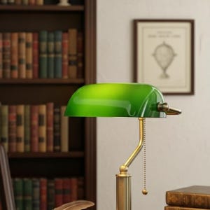 May include: A green-shaded banker's lamp with a brass base sits on a desk, next to an open book and a stack of books. A bookshelf filled with books is in the background. Two framed pictures hang on the wall.