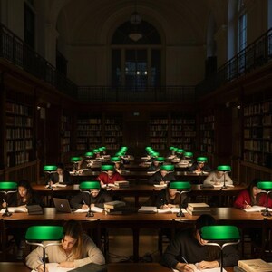 K&ouml;nnte beinhalten: Innenansicht einer Bibliothek mit Reihen von Schreibtischen und gr&uuml;nen Lampen. Studenten sitzen an den Schreibtischen, lernen und benutzen Laptops. B&uuml;cherregale s&auml;umen die W&auml;nde. Der Raum ist schwach beleuchtet und schafft eine konzentrierte Atmosph&auml;re.