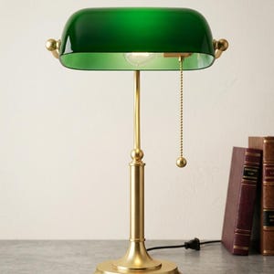 May include: A green glass banker's lamp with a brass base and accents. The lamp has a pull chain and is positioned on a gray surface next to several books. The lamp's shade is a deep, rich green.