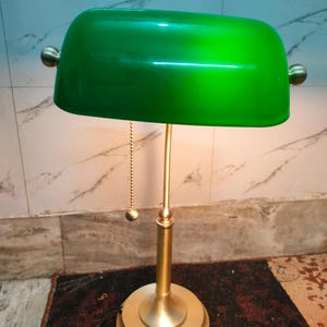May include: A green banker's lamp with a brass-colored base and stem. The lamp has a curved, rectangular shade and a pull chain. The base is round and tiered. The lamp is designed to provide focused light.