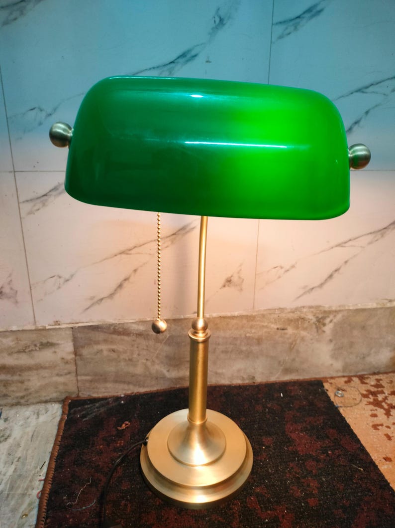 May include: A green banker's lamp with a brass-colored base and stem. The lamp has a curved, rectangular shade and a pull chain. The base is round and tiered. The lamp is designed to provide focused light.