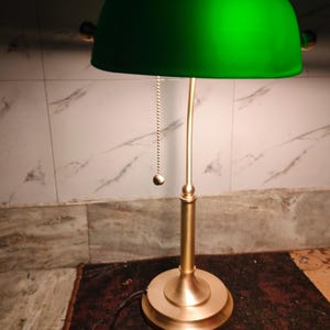 May include: A green-shaded desk lamp with a brass-colored base and stem. The lamp features a pull chain and a rounded shade. The lamp is on a dark red rug.