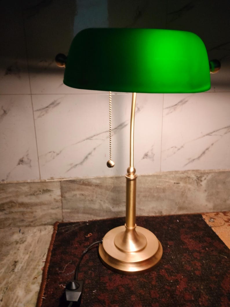 May include: A green-shaded desk lamp with a brass-colored base and stem. The lamp features a pull chain and a rounded shade. The lamp is on a dark red rug.