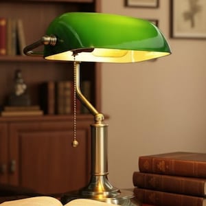 May include: A green-shaded desk lamp with a brass base and a pull chain sits on a desk next to an open book. The lamp illuminates the book and the surrounding area. Several books are stacked nearby.