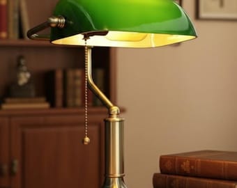 Könnte beinhalten: Eine grüne Schreibtischlampe mit Messingfuß und Zugkette steht auf einem Schreibtisch neben einem offenen Buch. Die Lampe beleuchtet das Buch und die Umgebung. Mehrere Bücher sind daneben gestapelt.