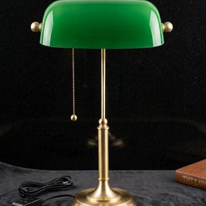 May include: A green-shaded desk lamp with a brass base and accents. The lamp features a pull chain and a cord with an on/off switch. The lamp's shade is a vibrant green, and the base is a warm brass color. The lamp is designed for desk use.