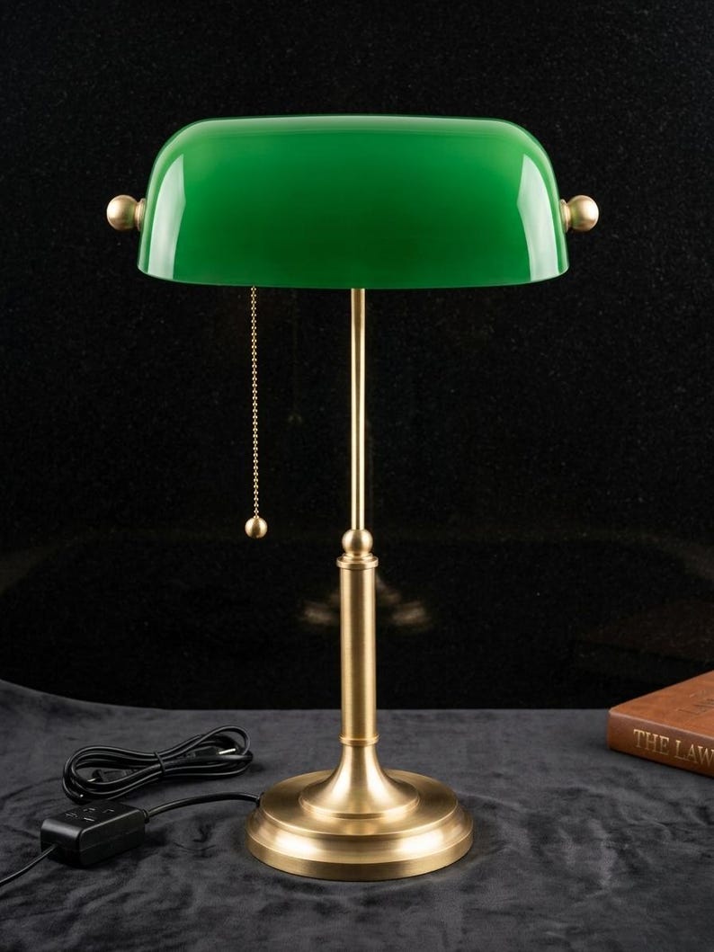 May include: A green-shaded desk lamp with a brass base and accents. The lamp features a pull chain and a cord with an on/off switch. The lamp's shade is a vibrant green, and the base is a warm brass color. The lamp is designed for desk use.