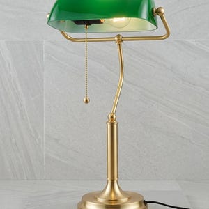 May include: A green-shaded desk lamp with a brass-colored base and frame. The lamp features a pull chain and a curved arm supporting the shade. The base is round, and the lamp is plugged into an outlet.