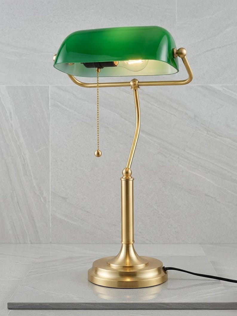 May include: A green-shaded desk lamp with a brass-colored base and frame. The lamp features a pull chain and a curved arm supporting the shade. The base is round, and the lamp is plugged into an outlet.