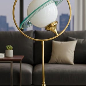 Puede incluir: Lámpara de mesa dorada única con una pantalla esférica de vidrio blanco rodeada por un anillo con rayas verdes, que se asemeja a un planeta. La lámpara tiene un tallo delgado y una base decorativa, creando una lámpara moderna y artística.