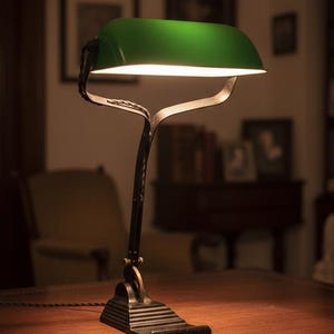 K&ouml;nnte beinhalten: Eine Schreibtischlampe im Vintage-Stil mit gr&uuml;nem Lampenschirm und schwarzem, verziertem Sockel. Die Lampe steht auf einem Holzschreibtisch und strahlt ein warmes Licht aus. Das Design erinnert an klassische Bankerlampen.