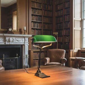K&ouml;nnte beinhalten: Eine Schreibtischlampe mit gr&uuml;nem Schirm, schwarzem Sockel und messingfarbenen Akzenten steht auf einem gro&szlig;en Holztisch. Die Lampe befindet sich in einer Bibliothek mit B&uuml;cherregalen, einem Kamin und einem Ledersessel.