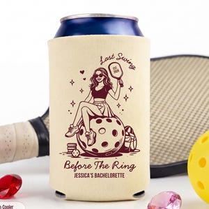 May include: Ivory-colored can cooler with a brown graphic of a woman with sunglasses holding a pickleball paddle, sitting on a pickleball. Text reads "Last Swing Before The Ring, Jessica's Bachelorette". A pickleball and paddle are in the background.
