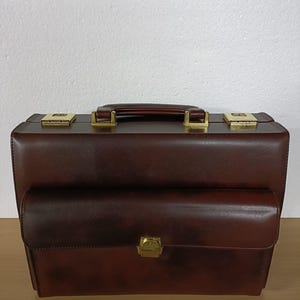 May include: A brown leather briefcase with a top handle and a front flap pocket. The briefcase features gold-coloured metal hardware, including clasps and a lock. The handle is a darker shade of brown than the body of the briefcase.