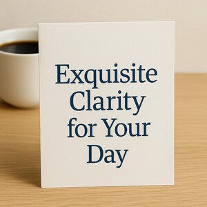 May include: A cream-colored card with the text "Exquisite Clarity for Your Day" in navy blue. A white mug filled with dark coffee is in the background, resting on a wooden surface.