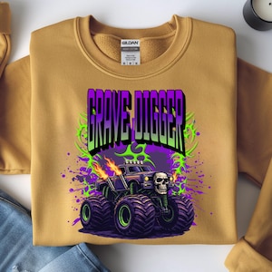 May include: Mustard-colored sweatshirt featuring a graphic of a monster truck with the text "GRAVE DIGGER". The monster truck is black with a skull on the front, flames, and purple and green accents. The sweatshirt has a crew neck and long sleeves.