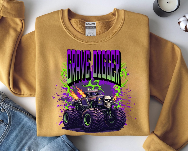 May include: Mustard-colored sweatshirt featuring a graphic of a monster truck with the text "GRAVE DIGGER". The monster truck is black with a skull on the front, flames, and purple and green accents. The sweatshirt has a crew neck and long sleeves.