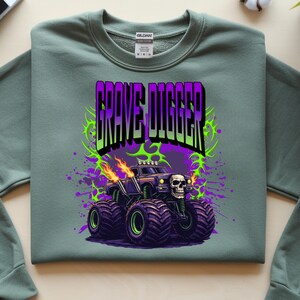 May include: Sage green sweatshirt featuring a graphic of a monster truck with the text "GRAVE DIGGER" in purple and green. The monster truck has large black tires with green rims, flames, and a skull on the front.