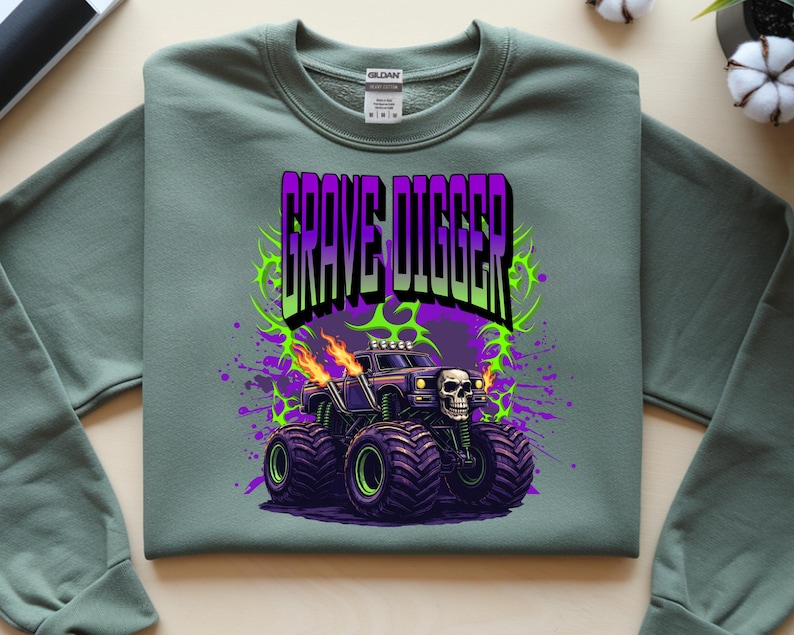 May include: Sage green sweatshirt featuring a graphic of a monster truck with the text "GRAVE DIGGER" in purple and green. The monster truck has large black tires with green rims, flames, and a skull on the front.