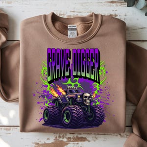 May include: A tan sweatshirt featuring a graphic of the monster truck Grave Digger. The design includes the text "Grave Digger" in purple and green, a skull, flames, and a purple and green splatter effect. The monster truck has large black tires.