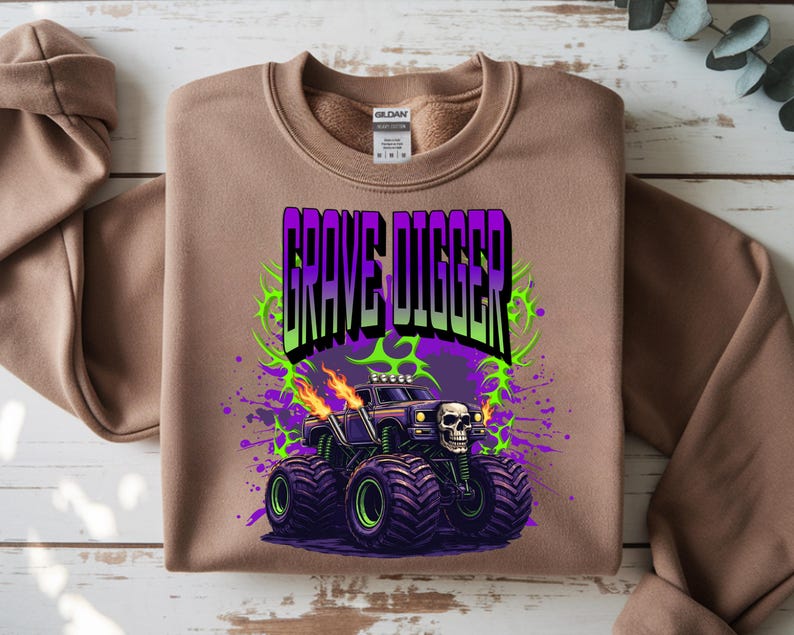 May include: A tan sweatshirt featuring a graphic of the monster truck Grave Digger. The design includes the text "Grave Digger" in purple and green, a skull, flames, and a purple and green splatter effect. The monster truck has large black tires.