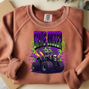 May include: A terracotta-colored sweatshirt featuring a graphic of a monster truck with the text "GRAVE DIGGER". The monster truck is black with purple and green accents, flames, and a skull.