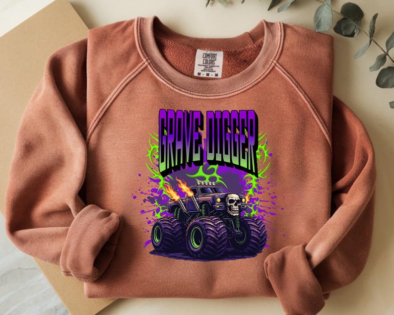 May include: A terracotta-colored sweatshirt featuring a graphic of a monster truck with the text "GRAVE DIGGER". The monster truck is black with purple and green accents, flames, and a skull.