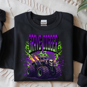 May include: Black sweatshirt featuring a graphic of a monster truck with the text "GRAVE DIGGER" in purple and green. The monster truck has a skull on the front and flames coming from the exhaust pipes. The design is surrounded by green and purple accents.