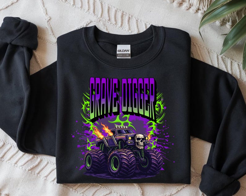 May include: Black sweatshirt featuring a graphic of a monster truck with the text "GRAVE DIGGER" in purple and green. The monster truck has a skull on the front and flames coming from the exhaust pipes. The design is surrounded by green and purple accents.