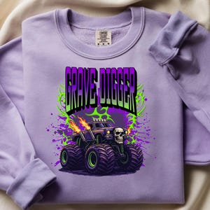 May include: A lilac-colored sweatshirt featuring a graphic of a monster truck with the text "GRAVE DIGGER". The monster truck has a skull on the front and flames coming from the exhaust pipes. The design includes green and purple accents.