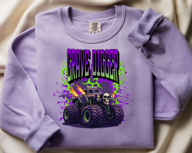 May include: A lilac-colored sweatshirt featuring a graphic of a monster truck with the text "GRAVE DIGGER". The monster truck has a skull on the front and flames coming from the exhaust pipes. The design includes green and purple accents.