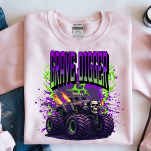 May include: A light pink sweatshirt featuring a graphic of a monster truck with the text "GRAVE DIGGER" in purple and green. The monster truck has a skull on the front and flames coming from the exhaust pipes. The design is set against a purple and green splatter background.