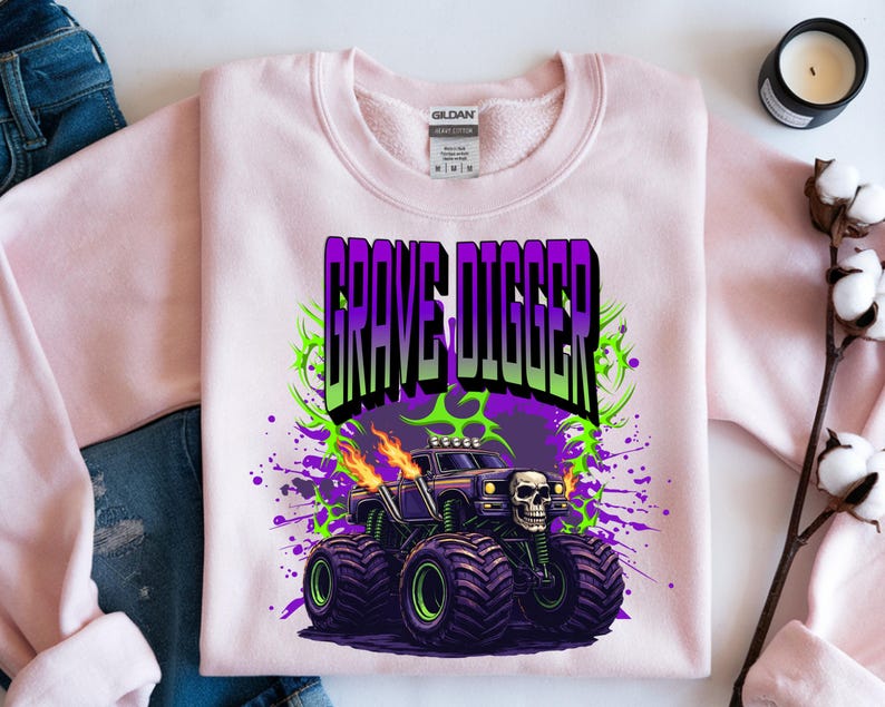 May include: A light pink sweatshirt featuring a graphic of a monster truck with the text "GRAVE DIGGER" in purple and green. The monster truck has a skull on the front and flames coming from the exhaust pipes. The design is set against a purple and green splatter background.