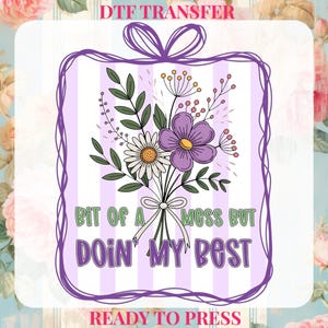 May include: DTF transfer featuring a floral bouquet with daisies and purple flowers, tied with a bow. The text "Bit of a mess but doin' my best" is displayed in green and purple. The background has vertical stripes.