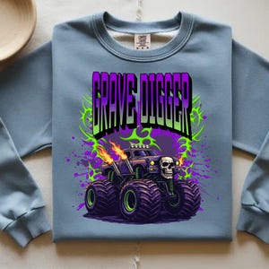 May include: A light blue sweatshirt featuring a graphic of a monster truck with the text "GRAVE DIGGER". The monster truck has a skull on the front and flames coming from the exhaust pipes. The design includes purple and green accents.