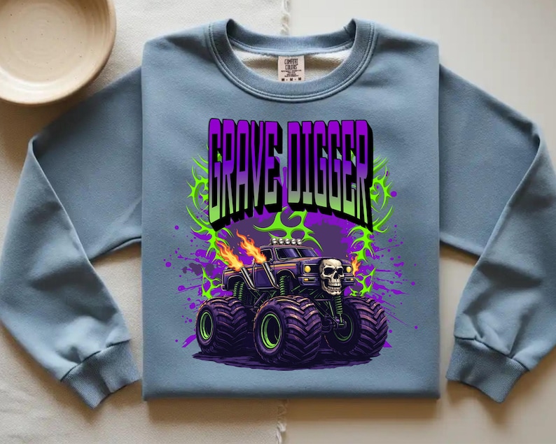 May include: A light blue sweatshirt featuring a graphic of a monster truck with the text "GRAVE DIGGER". The monster truck has a skull on the front and flames coming from the exhaust pipes. The design includes purple and green accents.