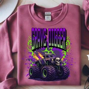 May include: A maroon sweatshirt featuring a graphic of a monster truck with the text "GRAVE DIGGER". The monster truck is black with green and purple accents, flames, and a skull. The design is surrounded by green and purple splatters.
