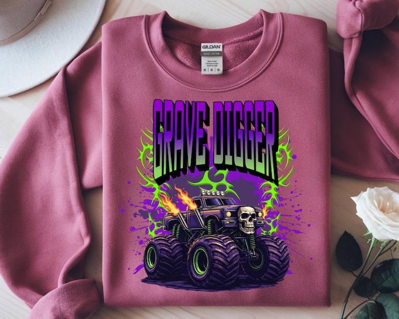 May include: A maroon sweatshirt featuring a graphic of a monster truck with the text "GRAVE DIGGER". The monster truck is black with green and purple accents, flames, and a skull. The design is surrounded by green and purple splatters.