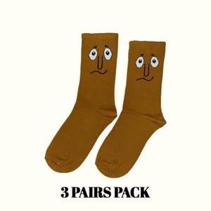 May include: Two pairs of mustard-colored socks with a cartoon face design. The socks have white eyes, a black nose, and a curved mouth. The text "3 PAIRS PACK" is printed in black at the bottom of the image.