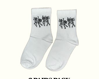 Puede incluir: Dos pares de calcetines blancos con un diseño de esqueleto negro. Los calcetines tienen un puño acanalado y un gráfico de esqueletos bailando. El texto "3 PAIRS PACK" está impreso en negro en la parte inferior de la imagen.