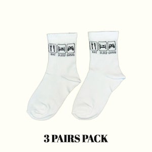 Peut inclure: Chaussettes de cheville blanches avec l'inscription "EAT SLEEP GAME" imprimée en noir au-dessus de la cheville. Le texte est accompagné de petites icônes noires d'une fourchette, d'un lit et d'une manette de jeu. L'inscription "3 PAIRS PACK" est imprimée en noir en bas.