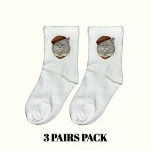 Op de afbeelding: Witte sokken met een grijze kat die een bruine baret en een overhemd met kraag draagt. De sokken hebben een geribbelde textuur en de tekst "3 PAIRS PACK" onderaan. De kattenillustratie is gecentreerd op elke sok.